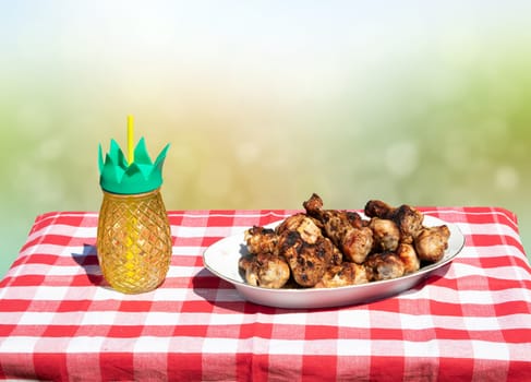 Fresh grilled chicken meat and a glass of juice on table with red checkered tablecloth and bright green white bokeh background. BBQ.
