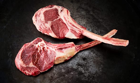 Two uncooked fatty ribeye or Delmonico steaks with the bone in viewed from above on an old textured blackened metal surface