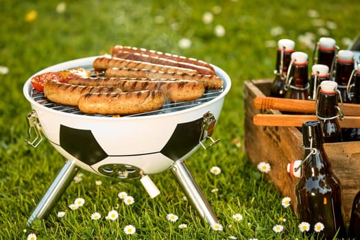 Sausages grilling on a soccer ball style BBQ alongside a wooden crate with bottles of craft beer in a spring meadow conceptual of the World Cup