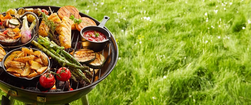Assortment of fresh healthy vegetables on a BBQ grilling over a hot fire in a green grassy spring or summer field in banner format with copy space