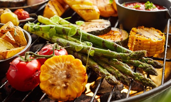 Healthy fresh summer vegetables grilling on a BBQ outdoors in the garden with zucchini, potato wedges, asparagus, tomato and corn on the cob garnished with herbs