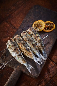 Fish barbecued on metal skewers, served on rustic cutting board on wooden table, viewed from high angle with copy space
