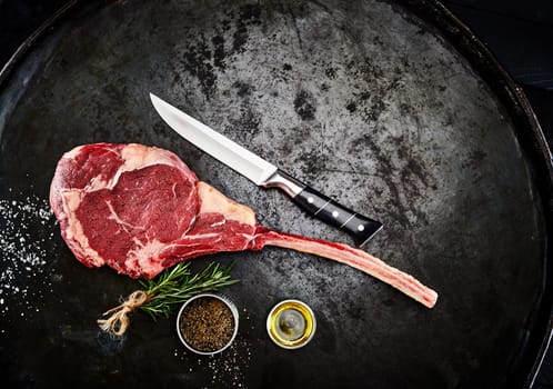 Preparing a ribeye or Delmonico steak in an overhead view of a tender fatty bone-in ribeye steak with spice rub, fresh rosemary and olive oil on a textured blackened metal pan
