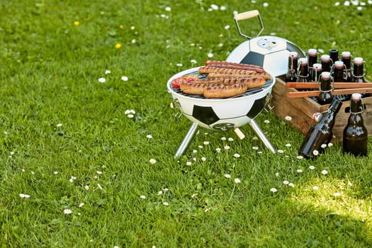 Celebration barbecue for the World Cup Soccer with sausages grilling over a ball shaped portable BBQ with a crate full of bottled beer alongside in a meadow