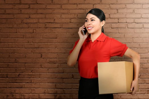 Beautiful asian delivery woman using mobile phone while bring the package with wall background