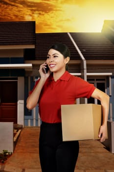Smiling asian courier woman with phone holding the parcel in front of house