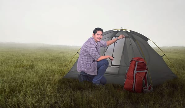 Happy asian tourist man with backpack prepare a tent on the meadow field