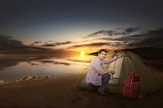 Smiling asian traveler man with backpack set up a tent beside the lake