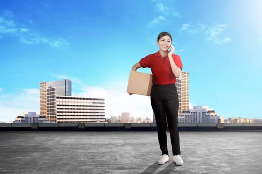 Young asian delivery woman with package talking on the cellphone on the city