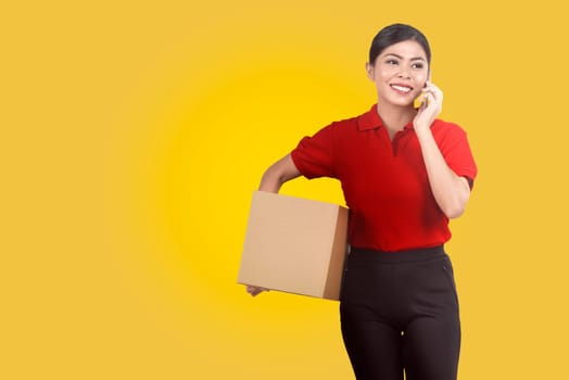 Pretty asian female courier using mobile phone while holding parcel over yellow background