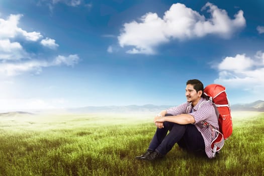 Smiling asian traveler man with backpack sitting and relaxing on the green field