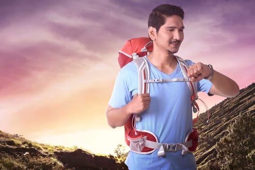 Smiling asian tourist with backpack checking his watch at mountain