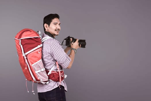 Young asian photographer with backpack holding camera over gray background