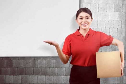 Young asian delivery woman carrying parcel and showing copyspace with whiteboard background
