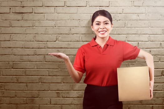 Beautiful asian delivery woman holding parcel and showing something over wall background