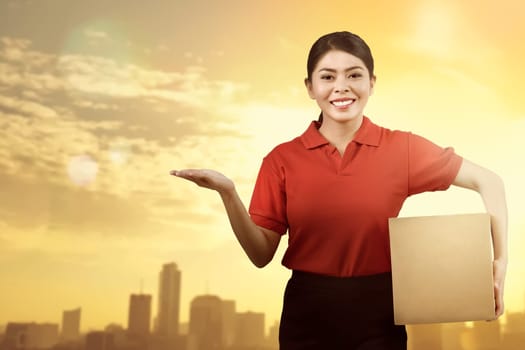 Cheerful asian woman courier with package and showing something over sunset background
