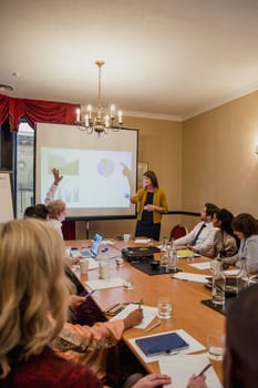 Rear view of a conference room filled with business people. One businessman is raising his hand to ask the presenter a question.