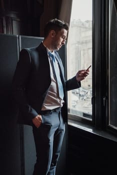 Side view of a young businessman standing next to a window in the office. He is checking his smart phone while waiting.
