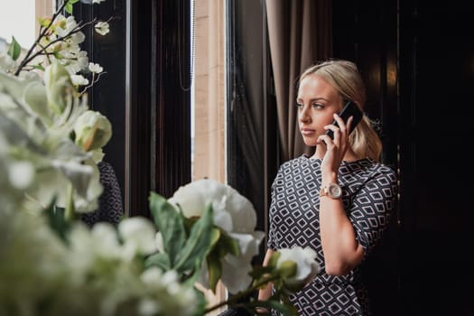Young adult businesswoman standing looking out the window. She is making an important phone call at work and has a serious facial expression.