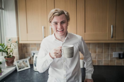 Front view of a young adult male standing in a kitchen looking at the camera with a cup of coffee.