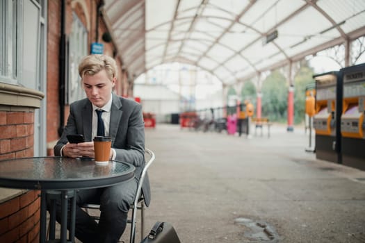 Young businessman sitting at a table in the train station. Using his smart phone to kill some time.