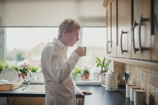 Side view of a young adult businessman standing at the kitchen counter blowing on a cup of tea to cool it down.