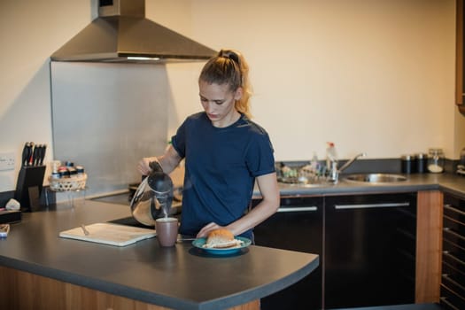 Front view of a young female adult pouring hot water into a cup for a cup of tea to have with her egg sandwich for breakfast.