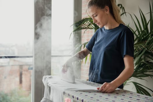 Side view of a young female adult ironing the laundry at home in the living room.