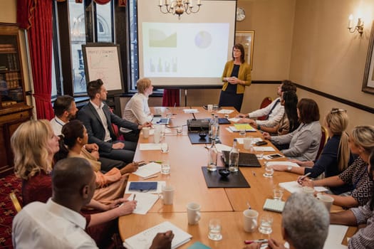 Mature businesswoman giving a presetation at a conference. Everyone at the meeting is sat around a table making notes and listening to the presentation.