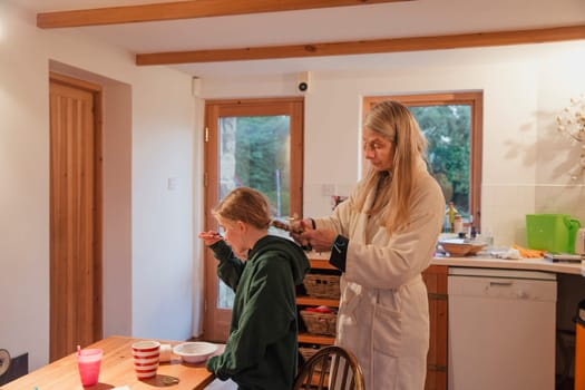Mother plaiting her daughters hair as she eats her breakfast in the kitchen.