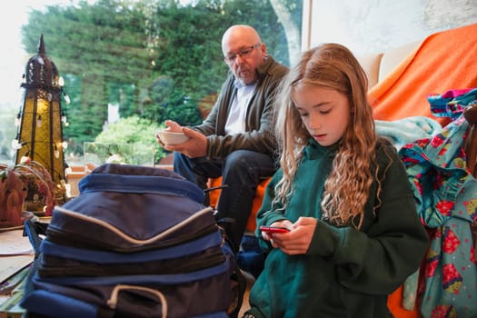 Little girl sitting on her mobile phone, her father is sat behind on the sofa watching over her shoulder.