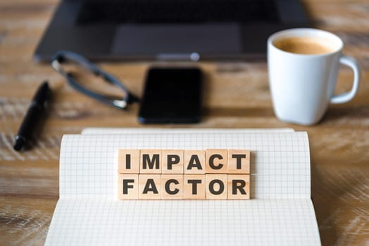 Closeup on notebook over vintage desk surface, front focus on wooden blocks with letters making Impact Factor text. Business concept image with office tools and coffee cup in background
