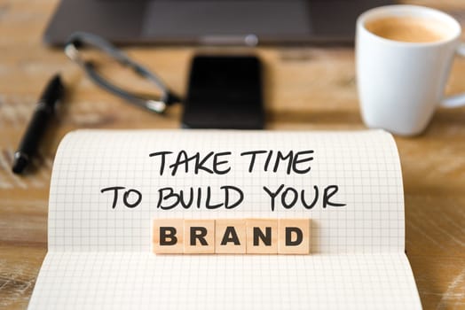 Closeup on notebook over vintage desk surface, front focus on wooden blocks with letters making Take Time To Build Your Brand text. Business concept image with office tools and coffee cup in background