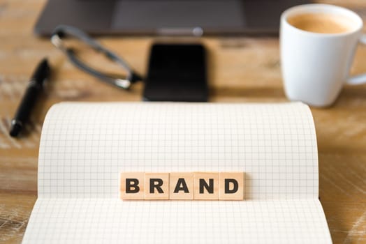 Closeup on notebook over vintage desk surface, front focus on wooden blocks with letters making Brand text. Business concept image with office tools and coffee cup in background