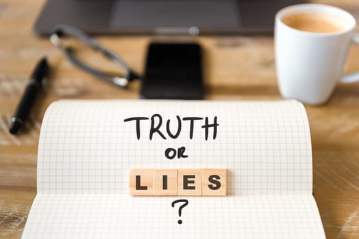 Closeup on notebook over vintage desk surface, front focus on wooden blocks with letters making Truth or Lies text. Business concept image with office tools and coffee cup in background