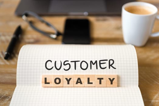 Closeup on notebook over vintage desk surface, front focus on wooden blocks with letters making Customer Loyalty text. Business concept image with office tools and coffee cup in background