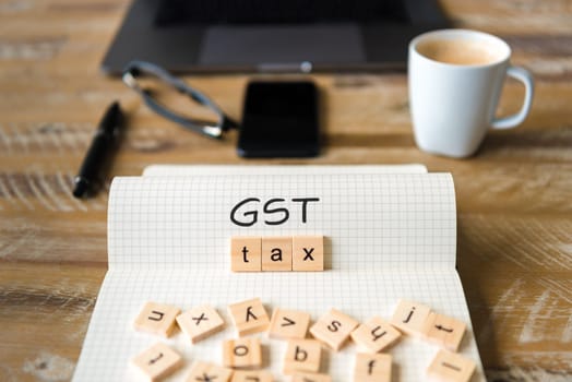 Closeup on notebook over vintage desk surface, front focus on wooden blocks with letters making GST Tax text. Business concept image with office tools and coffee cup in background