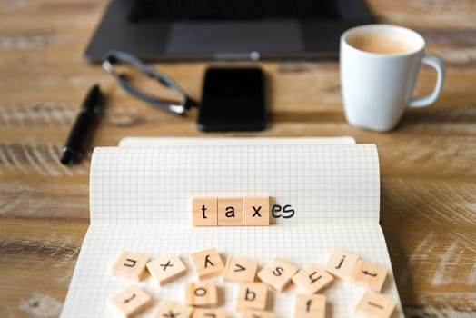 Closeup on notebook over vintage desk surface, front focus on wooden blocks with letters making Taxes text. Business concept image with office tools and coffee cup in background