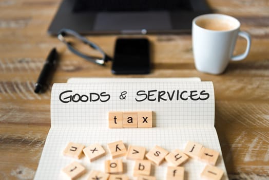 Closeup on notebook over vintage desk surface, front focus on wooden blocks with letters making Goods and Services Tax GST text. Business concept image with office tools and coffee cup in background