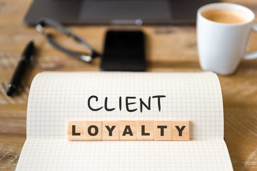 Closeup on notebook over vintage desk surface, front focus on wooden blocks with letters making Client Loyalty text. Business concept image with office tools and coffee cup in background