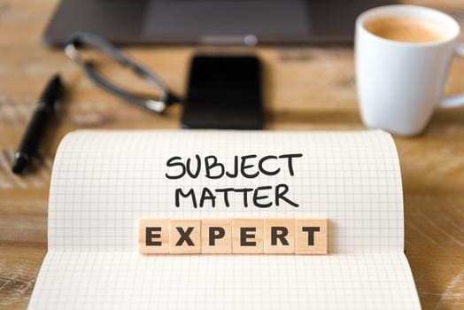 Closeup on notebook over vintage desk surface, front focus on wooden blocks with letters making Subject Matter Expert text. Business concept image with office tools and coffee cup in background