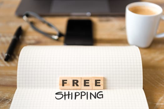 Closeup on notebook over vintage desk surface, front focus on wooden blocks with letters making Free Shipping text. Business concept image with office tools and coffee cup in background