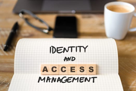 Closeup on notebook over vintage desk surface, front focus on wooden blocks with letters making Identity and Access Management. Business concept image with office tools and coffee cup in background