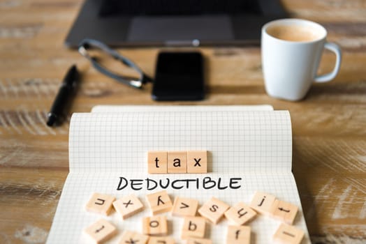 Closeup on notebook over vintage desk surface, front focus on wooden blocks with letters making Tax Deductible text. Business concept image with office tools and coffee cup in background