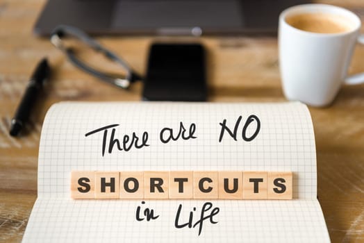 Closeup on notebook over vintage desk surface, front focus on wooden blocks with letters making There Are No Shortcuts In Life text. Business concept image with office tools and coffee cup in background