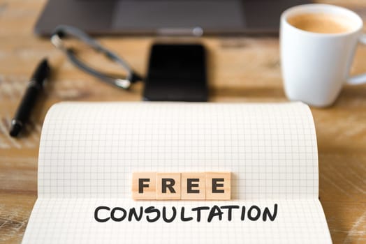 Closeup on notebook over vintage desk surface, front focus on wooden blocks with letters making Free Consultation text. Business concept image with office tools and coffee cup in background
