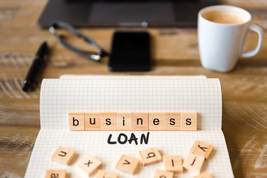 Closeup on notebook over vintage desk surface, front focus on wooden blocks with letters making Business Loan text. Business concept image with office tools and coffee cup in background