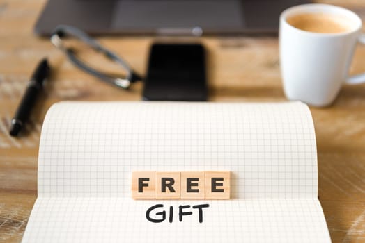 Closeup on notebook over vintage desk surface, front focus on wooden blocks with letters making Free Gift text. Business concept image with office tools and coffee cup in background