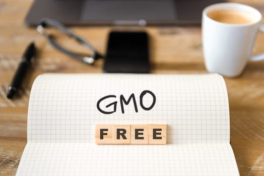 Closeup on notebook over vintage desk surface, front focus on wooden blocks with letters making GMO Free text. Business concept image with office tools and coffee cup in background