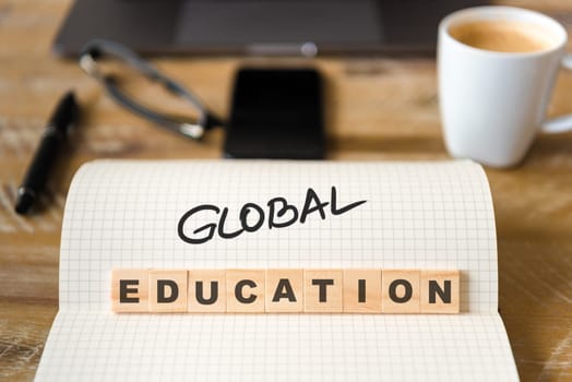 Closeup on notebook over vintage desk surface, front focus on wooden blocks with letters making Global Education text. Business concept image with office tools and coffee cup in background